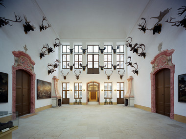 Ein weißer Saal, geziert von Jagdtrophäen an der Wand. Darunter befinden sich hölzerne große Türen mit einer roten Umrandung. Ebenso zu sehen sind zwei Kamine und eine Fensterfront.
