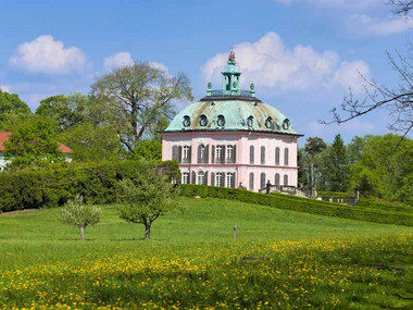 Auf einer grünen Wiese steht ein kleines Rokokoschlösschen in rosa mit einem mintgrünen Dach. 